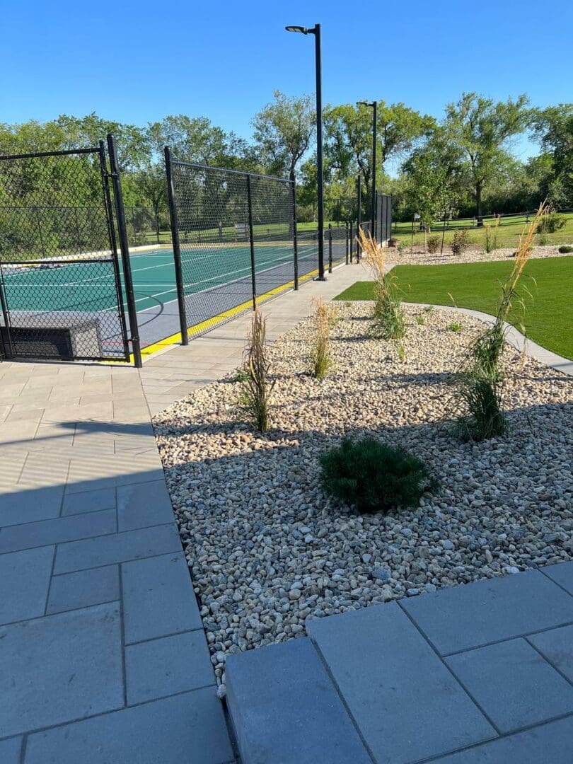 A tennis court with a green area and a paved walkway.